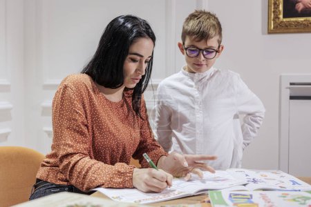 Photo for A dark-haired educator helps a blond boy with glasses do his homework in an elegant setting. - Royalty Free Image
