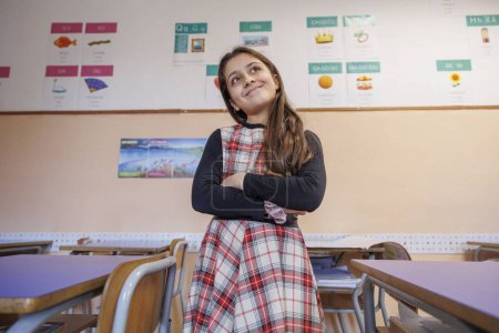 Foto de La alumna vestida con un vestido a cuadros está de pie con los brazos cruzados en un aula de una escuela - Imagen libre de derechos