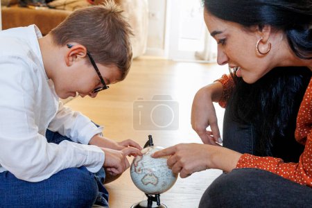 Téléchargez les photos : Fille adulte aux cheveux foncés montre un petit garçon un globe tandis qu'ils sont assis sur le sol à la maison - en image libre de droit