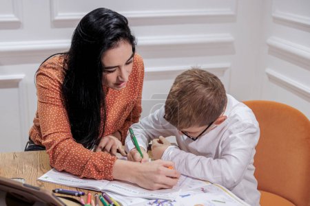 Foto de Un educador de cabello oscuro ayuda a un chico rubio con gafas a hacer sus deberes en un ambiente elegante. - Imagen libre de derechos