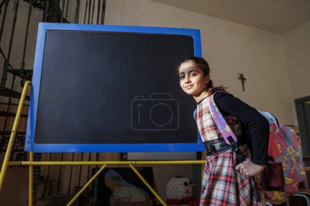 Téléchargez les photos : Heureuse écolière avec sac à dos près du tableau noir - en image libre de droit