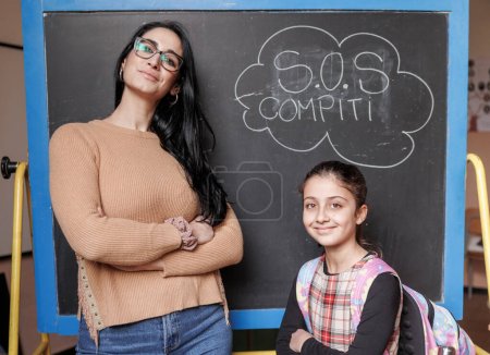 Photo for Teacher with her pupil look smiling, isolated on blackboard with written in Italian "sos Compiti" - Royalty Free Image
