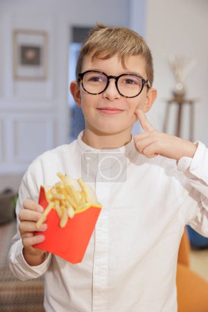 Photo for Blond boy holds french fries in his hand and shows that he likes them a lot. - Royalty Free Image