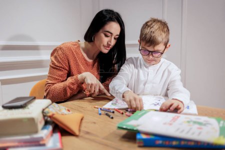 Foto de Un educador de cabello oscuro ayuda a un chico rubio con gafas a hacer sus deberes en un ambiente elegante. - Imagen libre de derechos