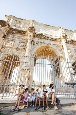 Foto de Madre con cinco niños turistas sentados bajo el espectacular Arco de Constantino, Roma, Italia. - Imagen libre de derechos