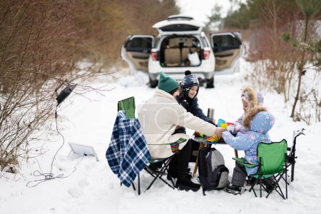Téléchargez les photos : Jeune femme avec des enfants dans la forêt d'hiver sur un pique-nique. Mère et trois enfants contre eux voiture avec coffre ouvert. - en image libre de droit