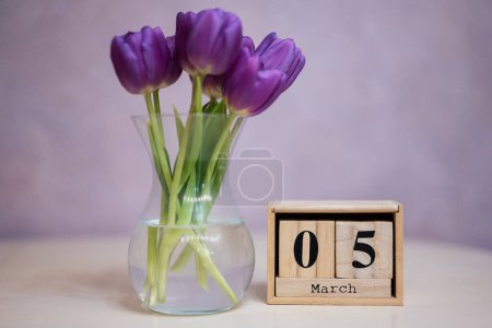 Hello spring! Wooden cubic calendar with date of 5 March surrounded with bouquet of purple tulips with green leaves in glass vase.