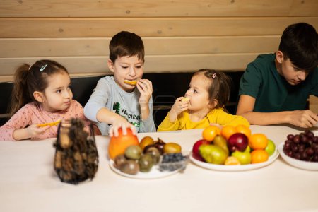 Téléchargez les photos : Quatre enfants mangent des fruits dans une maison de campagne en bois le week-end. - en image libre de droit