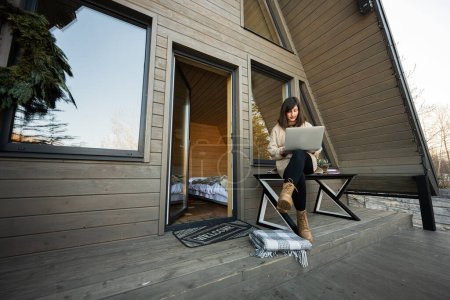 Photo for Remote work and escaping to nature concept. Woman works on laptop against tiny cabin house. - Royalty Free Image
