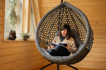 Photo for Remote work and escaping to nature concept. Woman sit in egg chair swing and read book in wooden tiny cabin house. - Royalty Free Image