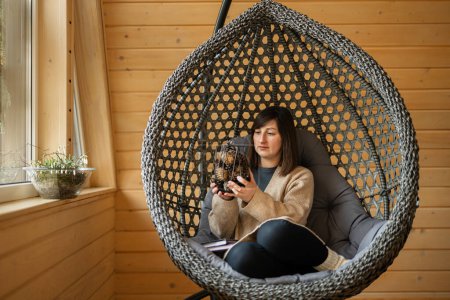 Photo for Remote work and escaping to nature concept. Woman sit in egg chair swing and holds a vase of cones in wooden tiny cabin house. - Royalty Free Image