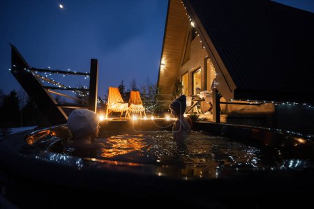 Foto de Los niños disfrutan bañándose en una bañera de hidromasaje de madera en la terraza de la casa. Bañera escandinava con chimenea para quemar madera y agua caliente. - Imagen libre de derechos
