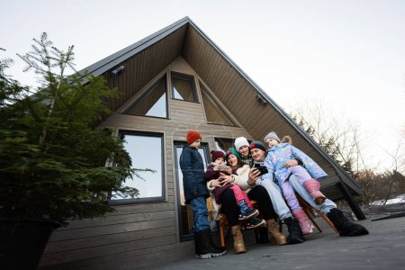 Téléchargez les photos : Grande famille avec quatre enfants assis sur la terrasse hors réseau petite maison dans les montagnes regardant les téléphones. - en image libre de droit