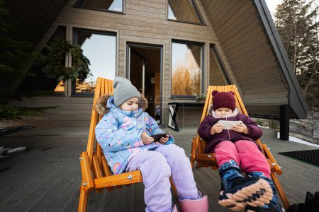 Zwei kleine Mädchen sitzen auf Stühlen auf der Terrasse vor dem kleinen Häuschen in den Bergen und gucken Zeichentrickfilme auf Mobiltelefonen.