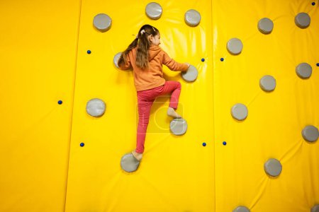 Little girl kid climbing wall at yellow playground park. Child in motion during active entertaiments.