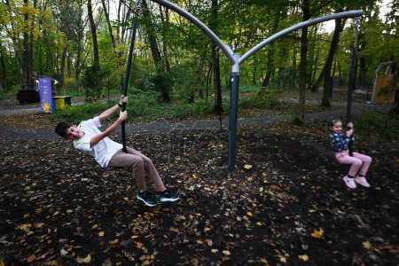 Téléchargez les photos : Garçon balançant sur une balançoire dans un parc d'automne avec sa petite sœur. - en image libre de droit