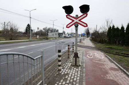 Escena urbana de carretera que muestra un cruce ferroviario con luces de señal, carril bici y barreras de seguridad en un día nublado.