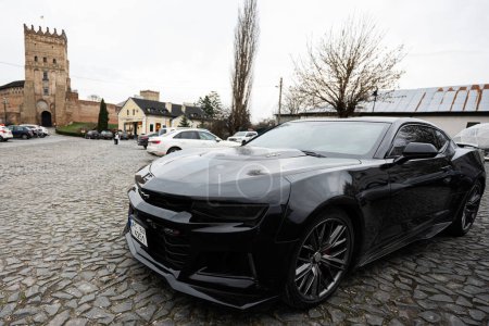 Photo for Lutsk, Ukraine - March, 2024: Black sports car Chevrolet Camaro ZL1 in focus, parked on a cobblestone square in front of an ancient fortified tower, portraying luxury and heritage. - Royalty Free Image