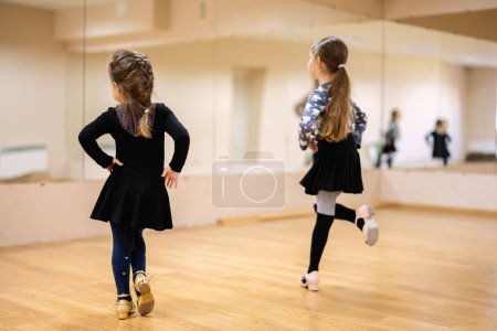 Two young girls practicing dance moves in a dance studio with mirrors. They are wearing dance attire and focusing on their reflection.