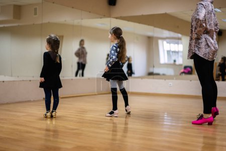 Two young girls in a dance studio, practicing with their instructor. They are dressed in dance attire, standing on a wooden floor with mirrors reflecting them.