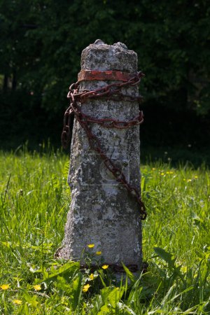 Photo for A lopsided gray cement post wrapped in grim rusty old chains stands alone on green grass in sunny summer weather. Forest in the background. Yellow flowers in the foreground. - Royalty Free Image