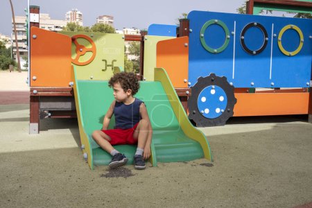 Lonely boy with curly hair sitting on a colorful slide.