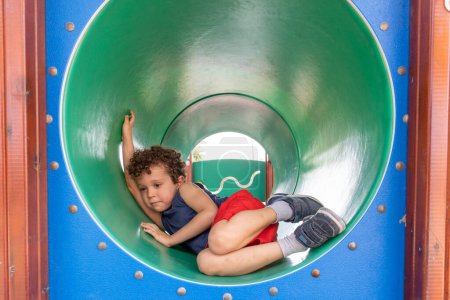 Curious boy with curly hair explores a green tunnel slide.