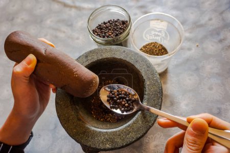 Téléchargez les photos : Épices dans la cuisine orientale exotique : un cuisinier montre une cuillère de poivre cru écrasé dans un mortier de pierre et pulvérisé au moyen d'un caillou - en image libre de droit
