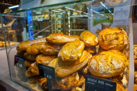 Téléchargez les photos : Pain bio français (boule de pain, pain de grain et baguette) dans la vitrine en verre d'une boulangerie dans un centre commercial en France, avec des étiquettes de prix indiquant la présence de gluten - en image libre de droit