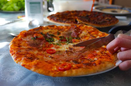 Photo for An appetizing Italian-styled pizza comprising cheese, prosciutto, tomato sauce and aromatic herbs is cut out in portions with scissors and served at the table for a lunch in France - Royalty Free Image