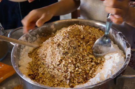 Photo for Artisanal making of a homemade cake in Southern Europe : a Frech female confectioner adds crushed almonds to dough and mixes the ingredients in a large metal bowl full of wheat flour with a spoon - Royalty Free Image
