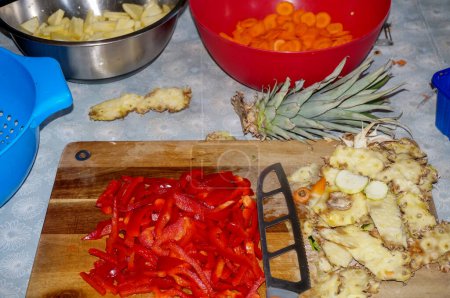 Photo for Artisanal preparation of a homemade mixed vegetable dish in a kitchen : fresh red peppers cut into slices, peelings of pineapple and a knife with hollow ground blade on a wooden chopping board - Royalty Free Image