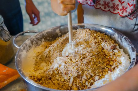 Photo for Artisanal making of a homemade cake in Southern Europe : a Frech female confectioner adds crushed almonds to dough and mixes the ingredients in a large mixing bowl full of wheat flour with a spoon - Royalty Free Image