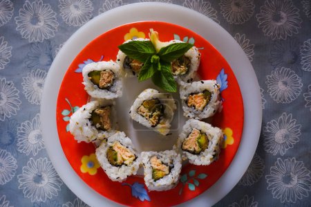 Téléchargez les photos : A few sushi-maki, a typical Japanese dish made of raw fish, rice and avocado, decorated with mint leaves ; the plate is colorful and lays on a blue, flowered table cloth - en image libre de droit