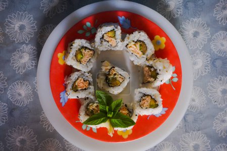 Photo for A few sushi-maki, a typical Japanese dish made of raw fish, rice and avocado, decorated with mint leaves ; the plate is colorful and lays on a blue, flowered table cloth - Royalty Free Image