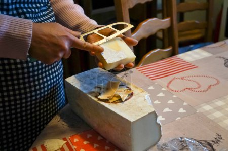 Foto de Aveyron, Francia - 1 de mayo de 2023 - Una ama de casa quita la corteza de un queso con un cuchillo de cocina, otro gran bloque de queso se coloca sobre la mesa; Aveyron rural produce queso de oveja o cabra famosa - Imagen libre de derechos