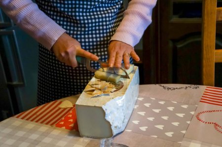 Téléchargez les photos : Aveyron, France - 1er mai 2023 - Une femme au foyer enlève la croûte d'un fromage avec un couteau de cuisine, un autre grand bloc de fromage est posé sur la table ; Aveyron rural produit célèbre fromage de brebis ou de chèvre - en image libre de droit