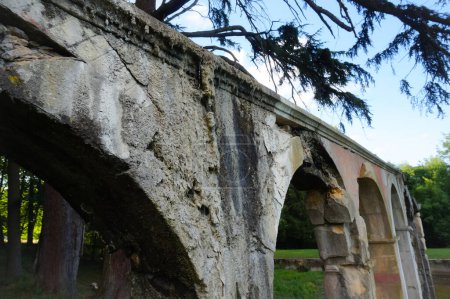 Téléchargez les photos : Remnants of an antique aqueduct in ruins, partly collapsed and damaged by moisture, facing an ancient pond partially filled with water, in the bucolic and wild atmosphere of a park in Rhne, France - en image libre de droit