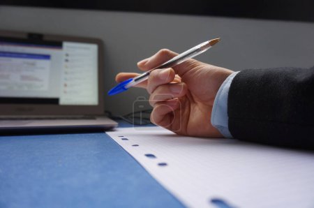 Téléchargez les photos : La main d'un homme sur un bureau, au-dessus d'une feuille de papier, tient un stylo à bille bleu, la personne porte une chemise bleue et un costume sombre, il y a un ordinateur personnel, avec l'écran sur - en image libre de droit