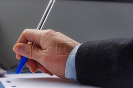 Téléchargez les photos : La main d'un homme sur un bureau, au-dessus d'une feuille de papier, tient un stylo à bille bleu - en image libre de droit