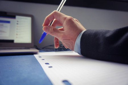 Foto de La mano de un hombre sobre un escritorio, encima de una hoja de papel, sostiene un bolígrafo azul, la persona lleva una camisa azul y un traje oscuro, hay una computadora personal, con la pantalla encendida - Imagen libre de derechos