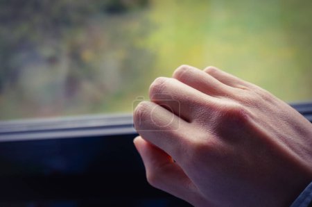 Photo for Closed right hand of a man, whose white skin is dehydrated and slightly damaged by the winter's cold ; the green grass of a park can be seen through the window in a blurred, bokeh background - Royalty Free Image