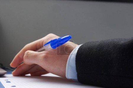 Foto de La mano de un hombre sobre un escritorio, encima de una hoja de papel, sostiene un bolígrafo azul - Imagen libre de derechos