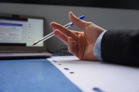 Téléchargez les photos : La main d'un homme sur un bureau, au-dessus d'une feuille de papier, tient un stylo à bille bleu, la personne porte une chemise bleue et un costume sombre, il y a un ordinateur personnel, avec l'écran sur - en image libre de droit