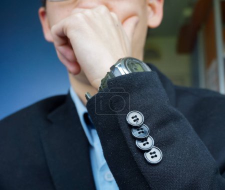 Foto de Un joven perplejo, sosteniendo su barbilla en su mano, está pensando en una actitud concentrada; lleva un traje negro con botones de manga, un reloj electrónico en la muñeca y una camisa azul. - Imagen libre de derechos