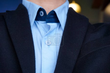 Téléchargez les photos : Détail des cols d'un costume noir et d'une chemise bleue, portés de manière décontractée, partiellement déboutonnés, par un jeune homme ; en détail, le tissu est légèrement endommagé, avec quelques fils de coton usagés et des peluches - en image libre de droit