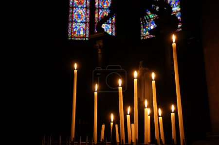 Foto de Reims, Francia - Marzo 2021 - Un conjunto de velas religiosas, brillando en el crepúsculo de una capilla lateral, con vidrieras de colores en el fondo, dentro de la nave de la Catedral de Notre-Dame - Imagen libre de derechos