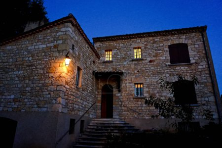 Téléchargez les photos : Photo de nuit d'une maison majestueuse dans le Tarn, dans le sud de la France, construite en calcaire dans le style ancien d'un château médiéval, avec escalier d'entrée avec une lanterne éclairée et fenêtres barrées éclairées - en image libre de droit
