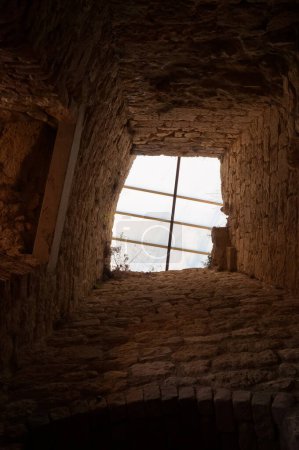 Foto de Castelnau-de-Lvis, Francia - Feb. 2020 - Vista de bajo ángulo de un pozo de luz dentro de las ruinas del castillo medieval, con paredes gruesas y una ventana de techo con barras, con césped silvestre creciendo en la piedra - Imagen libre de derechos