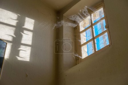 Photo for Low angle view of a huge and thick spider web on a muntin window having bars and illuminated by sunlight, inside an old and dusty house with light walls and high ceiling - Royalty Free Image
